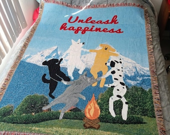 May include: A woven throw blanket with a light blue background featuring a mountain scene and dancing dogs. The phrase "Unleash happiness" is written in red. The blanket has a fringed edge with red and tan threads.