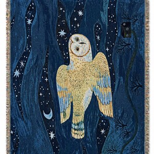 May include: A woven throw blanket featuring a large owl with outstretched wings against a dark blue night sky filled with stars and a crescent moon. The owl is primarily beige and yellow with blue accents. The blanket has a fringed edge.
