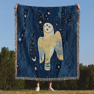 May include: A dark blue throw blanket featuring a large owl illustration with a night sky design. The owl is depicted in shades of yellow and blue, with a crescent moon and stars. The blanket has a fringed edge.