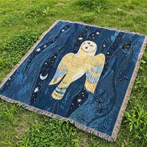May include: A woven throw blanket featuring a large owl design against a dark blue night sky with stars and a crescent moon. The owl is primarily yellow and tan with blue wing accents. The blanket has a fringed edge.