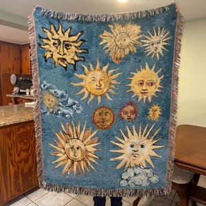 May include: A teal tapestry blanket featuring various sun designs with faces in shades of yellow, orange, and brown. The blanket has a fringed edge and is held up by a person.