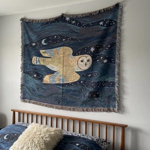 May include: A woven tapestry featuring a large owl in flight against a dark blue night sky with stars and a crescent moon. The owl is depicted in shades of beige and brown, with detailed feather patterns. The tapestry has a fringed edge.