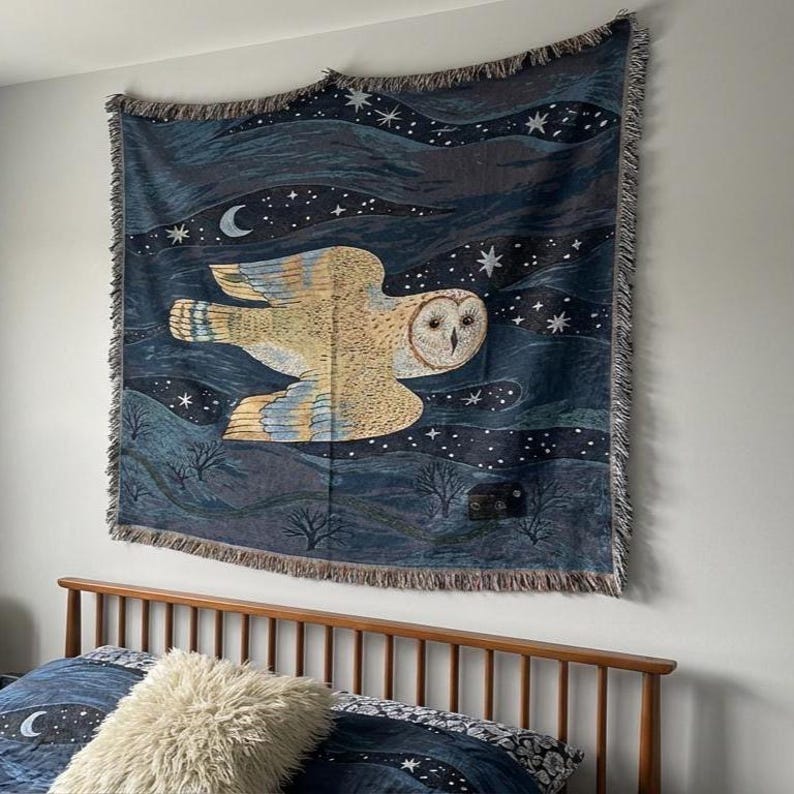 May include: A woven tapestry featuring a large owl in flight against a dark blue night sky with stars and a crescent moon. The owl is depicted in shades of beige and brown, with detailed feather patterns. The tapestry has a fringed edge.