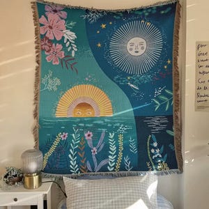 以下が含まれることがあります： 太陽と月をモチーフにした、織りのタペストリー。ターコイズブルーの背景に花と水の要素が描かれています。太陽は黄色とオレンジ色、月は白色です。縁はフリンジ加工が施されています。