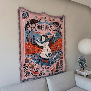 Puede incluir: Un tapiz rosa tejido con una ilustraci&oacute;n caprichosa. Muestra a una ni&ntilde;a con abrigo blanco, un gato negro y una escena de bosque. La palabra "CORALINE" est&aacute; en la parte superior y "SECRET FOREST" en la inferior. El tapiz tiene flecos.