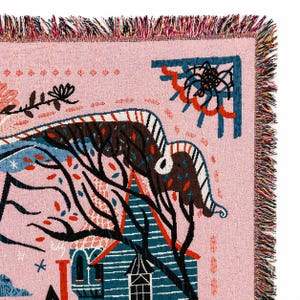 Puede incluir: Una manta tejida rosa con una ilustraci&oacute;n caprichosa. El dise&ntilde;o presenta una casa, &aacute;rboles y una telara&ntilde;a en tonos de azul, marr&oacute;n y rojo. La manta tiene un borde con flecos.
