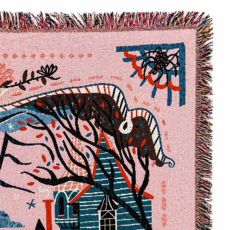 Puede incluir: Una manta tejida rosa con una ilustraci&oacute;n caprichosa. El dise&ntilde;o presenta una casa, &aacute;rboles y una telara&ntilde;a en tonos de azul, marr&oacute;n y rojo. La manta tiene un borde con flecos.