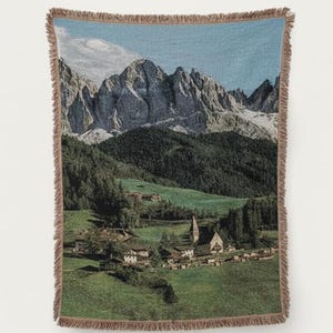 Puede incluir: Una manta tejida que representa un paisaje de montaña con un pueblo y una iglesia. La manta tiene un borde con flecos y presenta un valle verde, un cielo azul y una cordillera. Esta manta decorativa es un accesorio para el hogar.