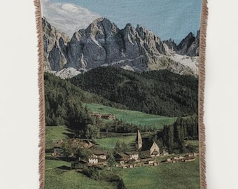 Può includere: Una coperta tessuta raffigurante un paesaggio montano con un villaggio e una chiesa. La coperta ha un bordo con frange e presenta una valle verde, un cielo blu e una catena montuosa. Questa coperta decorativa è un accessorio per la casa.