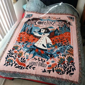 Puede incluir: Una manta tejida rosa con una ilustraci&oacute;n caprichosa. El dise&ntilde;o incluye una ni&ntilde;a con abrigo blanco, un gato negro y la frase "Secret Forest". Tambi&eacute;n incluye la palabra "Coraline". Tiene bordes con flecos.