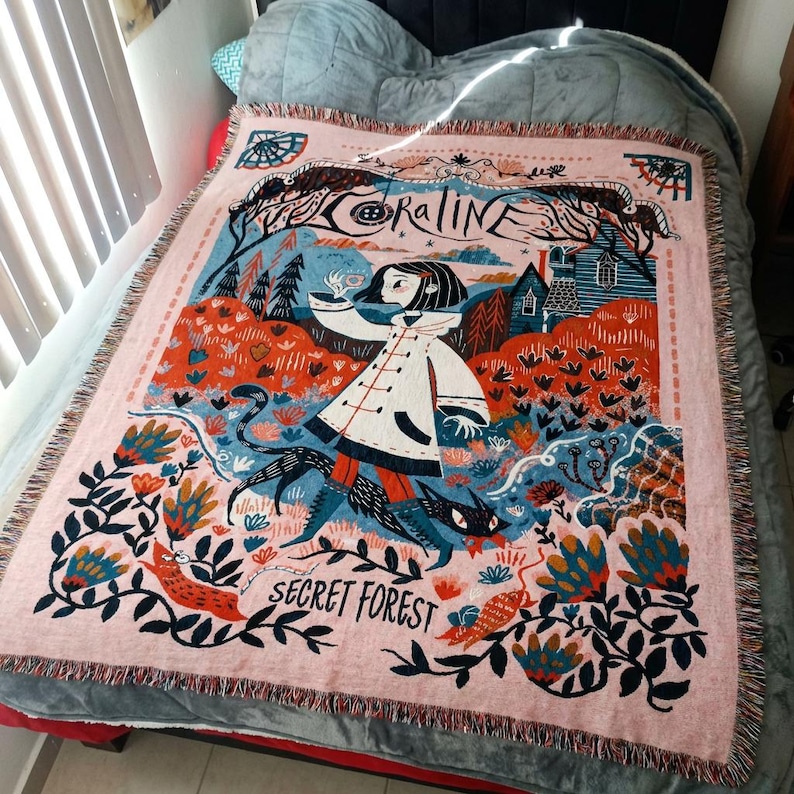 Puede incluir: Una manta tejida rosa con una ilustraci&oacute;n caprichosa. El dise&ntilde;o incluye una ni&ntilde;a con abrigo blanco, un gato negro y la frase "Secret Forest". Tambi&eacute;n incluye la palabra "Coraline". Tiene bordes con flecos.