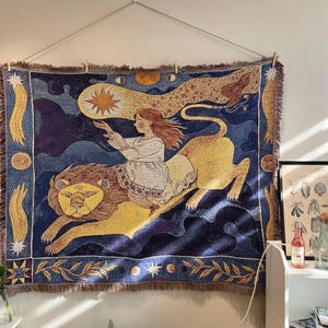 Peut inclure: Une tapisserie tissée représentant une scène céleste avec une femme chevauchant un lion. Le design utilise des teintes de bleu, de jaune et de blanc, et a une bordure à franges. La femme tend la main vers une étoile.