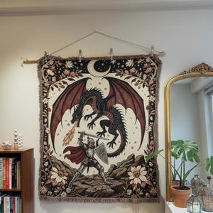 Peut inclure: Une tapisserie représentant un chevalier combattant un dragon. Le dragon est noir avec des ailes rouges, et le chevalier est en armure argentée. Le fond est crème avec des motifs floraux et célestes, et la tapisserie est frangée.