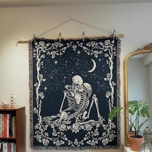 May include: A dark blue woven tapestry featuring a skeleton couple embracing under a crescent moon and stars. The design is framed by a floral border with roses and vines. The tapestry is hung on a wooden rod.