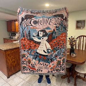 Puede incluir: Una manta tejida con una colorida ilustraci&oacute;n de la pel&iacute;cula "Coraline". El dise&ntilde;o incluye una ni&ntilde;a con un abrigo blanco, una casa y las palabras "Coraline" y "Secret Forest". La manta tiene un borde con flecos.