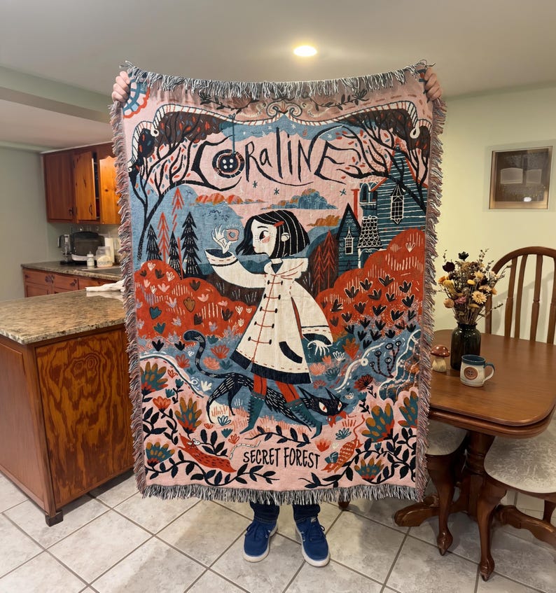 Puede incluir: Una manta tejida con una colorida ilustraci&oacute;n de la pel&iacute;cula "Coraline". El dise&ntilde;o incluye una ni&ntilde;a con un abrigo blanco, una casa y las palabras "Coraline" y "Secret Forest". La manta tiene un borde con flecos.