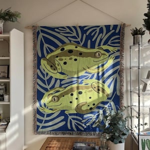 May include: A woven wall hanging featuring two stylized green frogs with brown spots against a blue background with white leaf patterns. The tapestry is rectangular with a fringed edge and is suspended from a wooden rod.