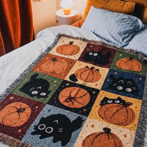 Op de afbeelding: Een geweven plaid met een rasterpatroon van afwisselende vierkanten. De vierkanten tonen oranje pompoenen en zwarte katten tegen een achtergrond van crème, groen, donkerblauw, geel en paars, geaccentueerd met kleine gele sterretjes.