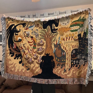 May include: Woven throw blanket with a detailed illustration depicting a mythological scene. The design features figures in boats, dragons, and a tree, all in shades of tan, black, and brown. The blanket has a fringed edge.