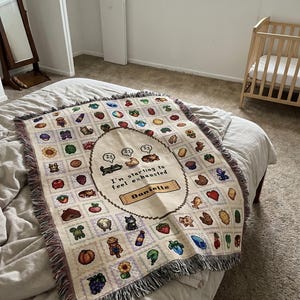 Op de afbeelding: Een cr&egrave;mekleurige sprei met een herhalend patroon van gepixelde afbeeldingen van verschillende objecten en dieren. Het midden bevat de tekst "I'm starting to feel exhausted" en de naam "Danielle". De sprei heeft een gefranjerde rand.