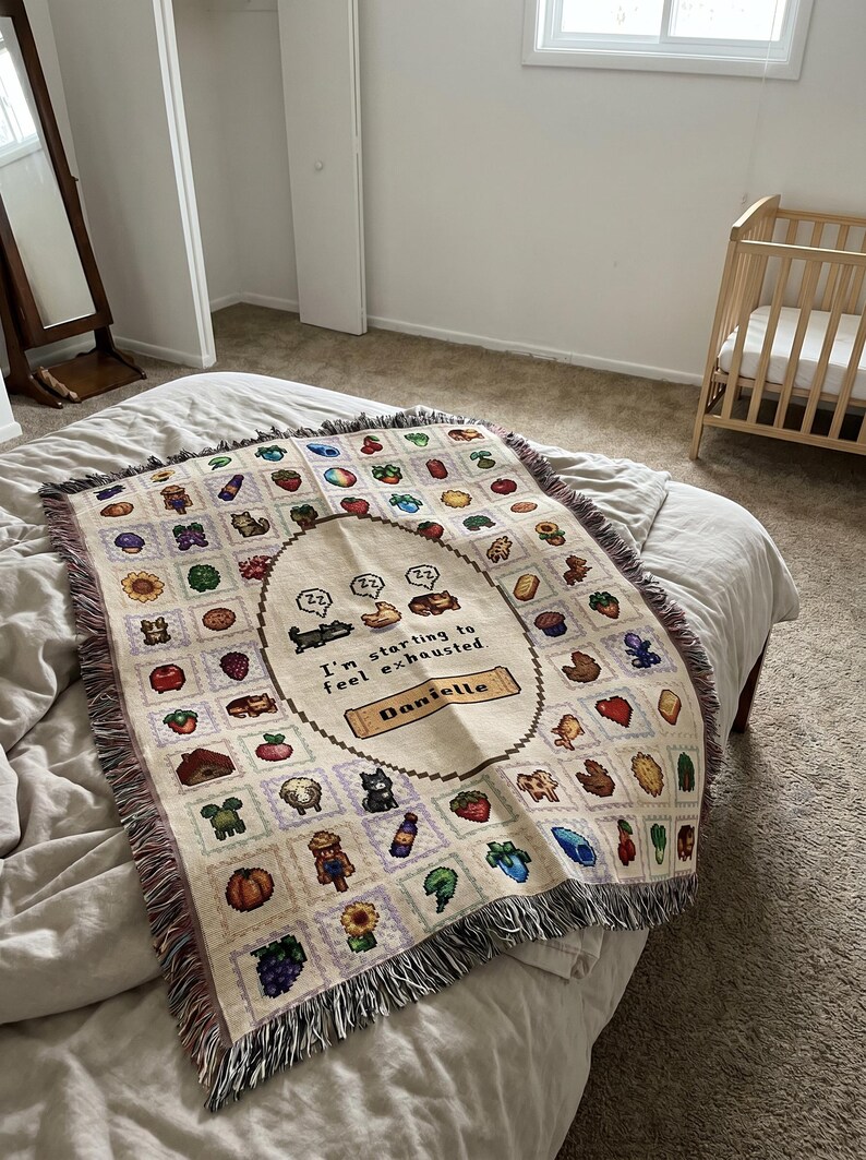 Op de afbeelding: Een cr&egrave;mekleurige sprei met een herhalend patroon van gepixelde afbeeldingen van verschillende objecten en dieren. Het midden bevat de tekst "I'm starting to feel exhausted" en de naam "Danielle". De sprei heeft een gefranjerde rand.