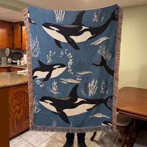 May include: A woven throw blanket featuring a repeating pattern of black and white orca whales swimming against a blue background. The design includes white illustrations of seaweed and bubbles, finished with a decorative fringed edge on all sides.