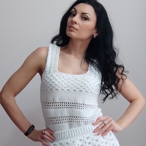 Puede incluir: Un top blanco de crochet con escote cuadrado y un delicado patrón de calado. El top es sin mangas y tiene un corpiño ajustado.