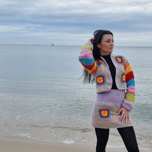 Puede incluir: Un conjunto de top y falda de crochet morado con diseños cuadrados coloridos. El top tiene rayas arcoíris en las mangas. La falda es más corta y se lleva con medias negras.