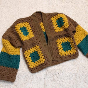 Può includere: Un cardigan marrone all'uncinetto con motivi quadrati gialli e turchesi. Le maniche presentano strisce alternate marroni, gialle e turchesi. Capo fatto a mano con design aperto.
