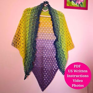 May include: A handmade crochet shawl in a gradient of colors, including yellow, green, and purple. The open weave design is visible, and the shawl is hanging on a hanger. The image also includes a pink circle with the text "PDF US Written Instructions Video Photos".