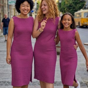 May include: Three sleeveless, magenta-colored crochet dresses. The dresses have a boat neck and a slit on the side. The women are smiling and standing on a city street. The dresses appear to be handmade.