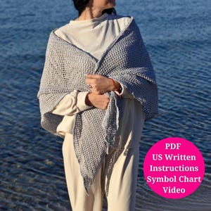 Puede incluir: Un chal de crochet gris con un delicado patrón de calado. El chal está drapeado sobre una persona que lleva una camisa blanca y pantalones beige. La imagen incluye el texto "PDF US Written Instructions Symbol Chart Video".