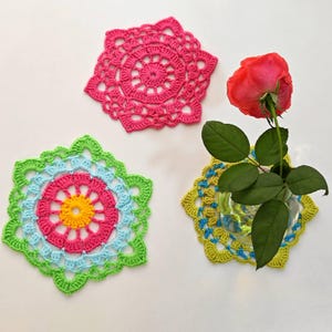 May include: Three crocheted doilies in pink, green, blue, and yellow. The doilies are circular and have a flower-like design. A single pink rose is in a glass vase on top of one of the doilies.