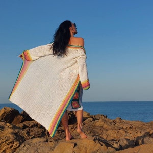 May include: A white crochet cardigan with a rainbow stripe detail. The cardigan is open and flowing, with a relaxed fit. The model is standing on a rocky beach, facing away from the camera.