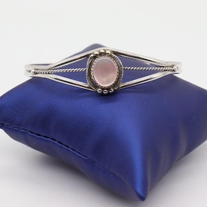 May include: A silver-toned cuff bracelet with a central oval-shaped pink gemstone. The bracelet features a twisted rope design and is displayed on a blue pillow.
