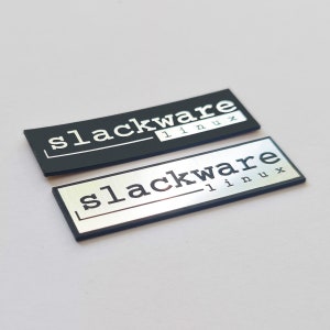 Peut inclure: Deux badges rectangulaires en métal. L'un est noir avec le texte blanc "slackware linux". L'autre est argenté avec le texte noir "slackware linux". Les deux ont une fine bordure.