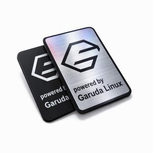 Puede incluir: Dos insignias rectangulares de metal. Una es negra con un diseño geométrico blanco y el texto "powered by Garuda". La otra es plateada con el mismo diseño y texto. También está presente el texto "powered by Garuda Linux".