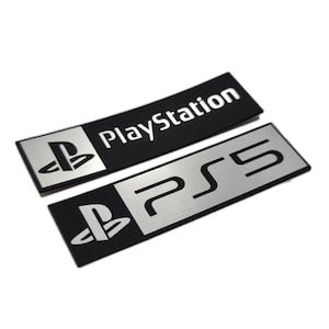 Puede incluir: Dos etiquetas rectangulares de metal con el logotipo de PlayStation. La etiqueta superior es negra con texto blanco que dice "PlayStation". La etiqueta inferior es plateada con texto negro que dice "PS5".