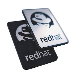Op de afbeelding: Twee metalen badges met het Red Hat-logo en het woord "redhat" in zwarte letters. De bovenste badge is zilverkleurig met een geborstelde afwerking en de onderste badge is zwart.