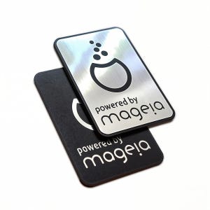 Op de afbeelding: Twee metalen labels, een zilveren en een zwarte, met een logo en de tekst "powered by magela!"
