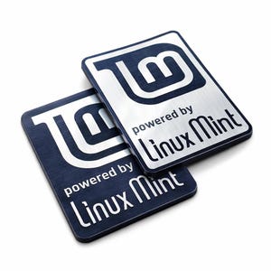 Op de afbeelding: Twee rechthoekige metalen stickers met een geborstelde zilveren afwerking en donkerblauwe accenten. Elke sticker toont het Linux Mint-logo bovenaan en de tekst "powered by Linux Mint" in een strak, schreefloos lettertype eronder.