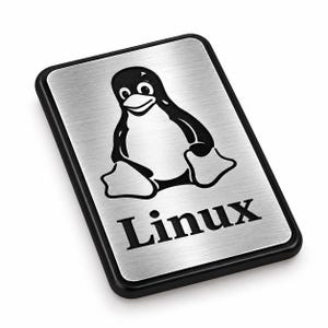 Op de afbeelding: Een rechthoekig plaatje met een geborstelde zilverkleurige metalen afwerking en een dikke zwarte rand. Het midden toont een zwarte afbeelding van een zittende pinguïn, geplaatst boven het woord Linux in een vetgedrukt, zwart schreeflettertype.