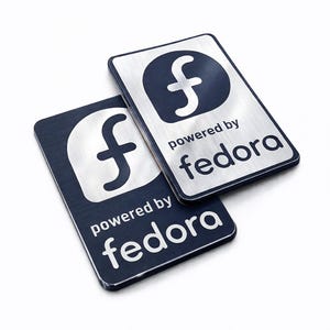 Puede incluir: Dos placas rectangulares de metal con acabado plateado cepillado y detalles en azul oscuro. Cada placa presenta una letra f minúscula estilizada dentro de un círculo azul oscuro, con el texto "powered by fedora" impreso en azul oscuro.