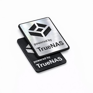 Könnte beinhalten: Zwei rechteckige TrueNAS-Abzeichen. Eines ist silberfarben mit schwarzem Logo und Text, das andere ist schwarz mit weißem Text. Beide Abzeichen tragen den Text "powered by TrueNAS". Das Logo ist ein stilisierter Würfel.