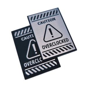 Puede incluir: Dos señales de advertencia con bordes rayados en blanco y negro. La señal superior es plateada con el texto "CAUTION OVERCLOCKED" y un signo de exclamación negro en un triángulo. La señal inferior es negra con el texto "CAUTION OVERCLOCKED" y un signo de exclamación blanco en un triángulo.