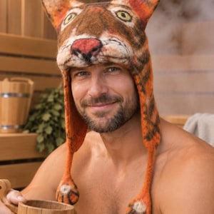 May include: A felted wool hat shaped like a tiger head with pointed ears, a pink nose, and wide eyes. The hat features long side flaps that hang down, each ending in a small, round, tiger-paw-shaped tassel with black stripes and white accents.