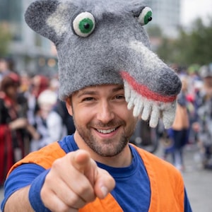 Puede incluir: Una persona con un sombrero de lobo gris con ojos verdes, dientes blancos y boca roja. La persona también lleva una camisa azul y un chaleco naranja, señalando al espectador.