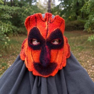 May include: A vibrant orange and red felted mask featuring a textured, organic shape with deep black accents around the eye openings, nose, and mouth area. The mask has a scalloped top edge and a structured, layered appearance with visible fiber details.