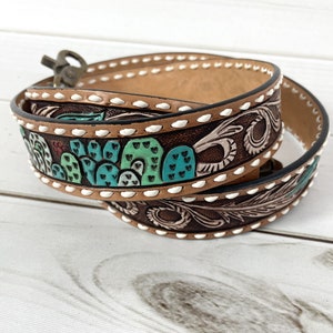 May include: A brown leather belt with a white stitching detail and a cactus design. The belt is decorated with green, blue, and red paint.