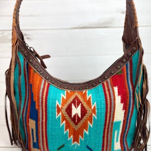 May include: A turquoise, orange, red, and white patterned hobo bag with brown leather trim and fringe. The bag has a zipper closure and a top handle.